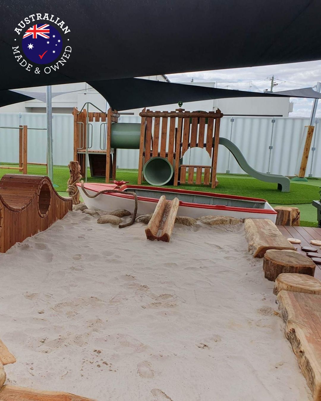 Outdoor play space featuring a timber preschool combination unit with slide and tunnel, surrounded by natural log elements, sandpit, and boat-themed play structure. Australian-made and owned.