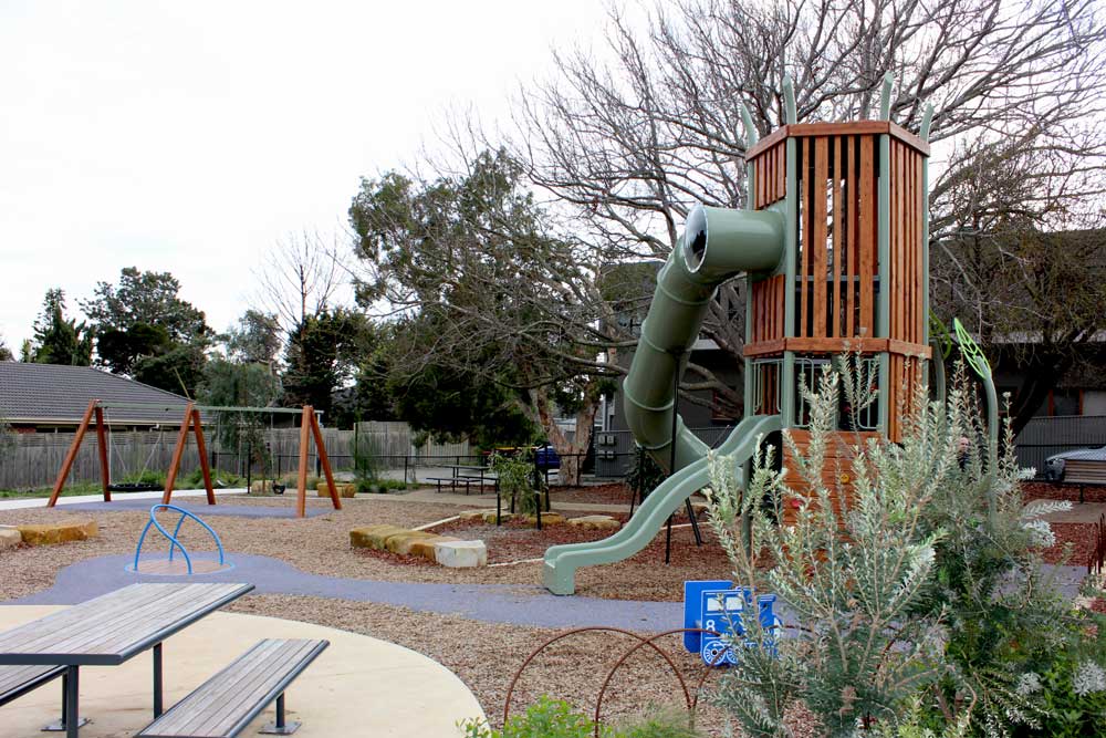 A nature-themed playground at Kareela Road Reserve in Frankston, designed and installed for Frankston City Council. It features a tall timber and steel play tower with enclosed spiral slide and straight slide, swings, a blue climbing spinner, picnic tables, and natural landscaping with plants, boulders, and mulch pathways.