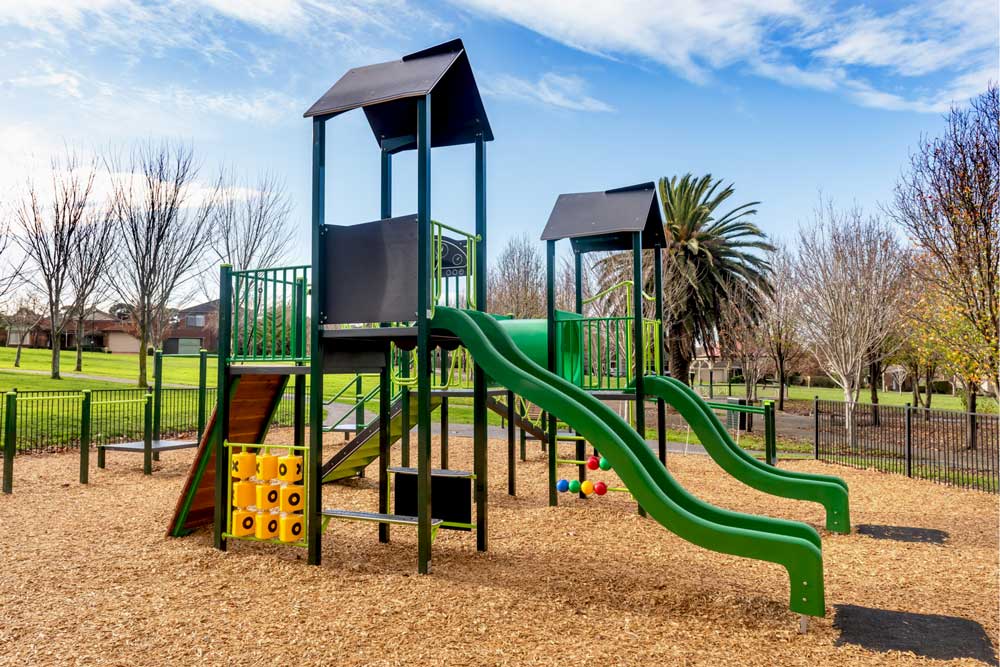 Front view of the Quokka Combination Unit at Village Green, Beaconsfield, highlighting two slides, climbing features, and interactive panels beneath the structure. The surrounding park provides open green space and seating for families.