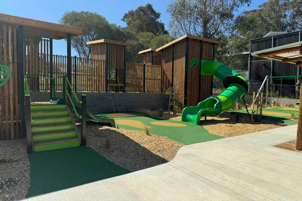 Custom playground installation in Orbost showing timber cladding towers connected by platforms, green spiral slide, low steps and climbing net on safety surfacing